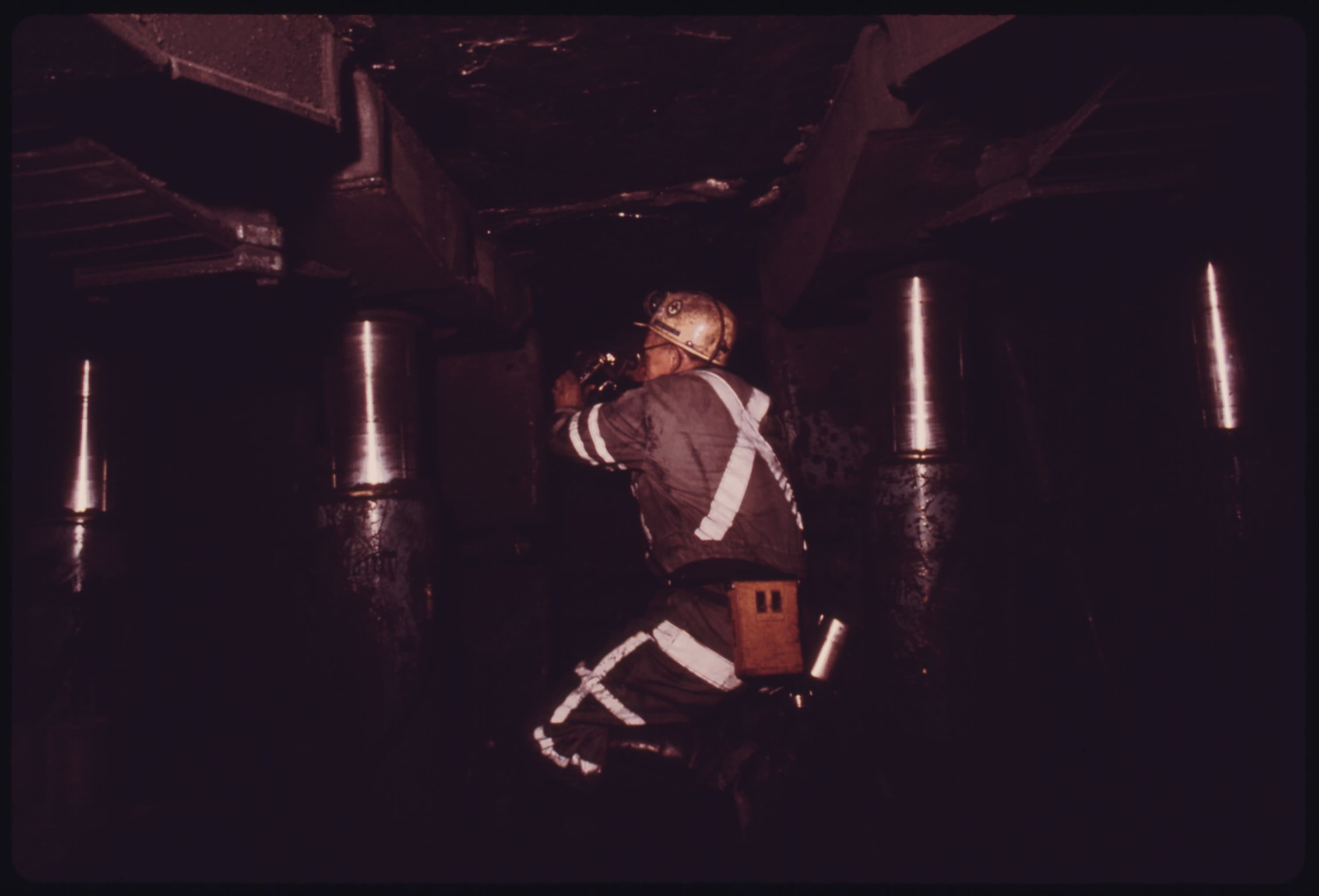 A federal coal mine inspector examining underground conditions among longwall support jacks, wearing a hard hat and high-visibility gear