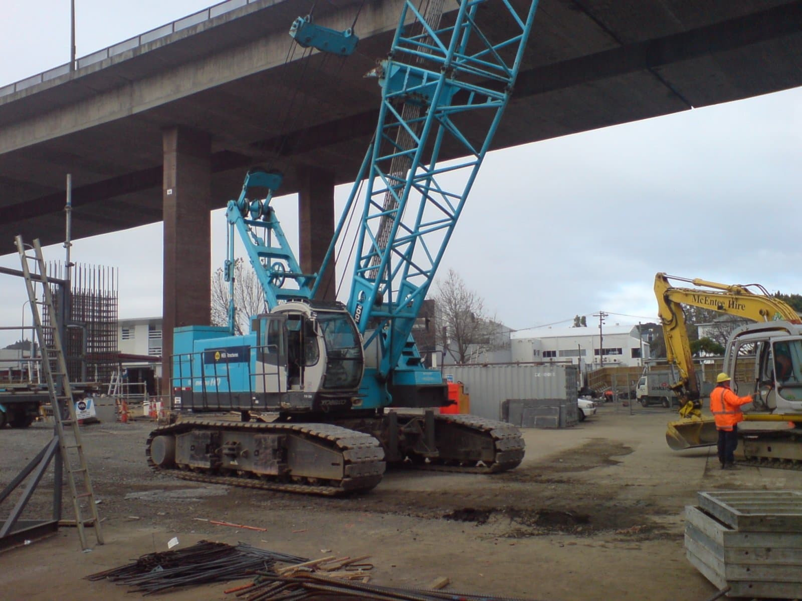 A large crawler crane and a tracked excavator on a bridge construction site, with concrete piers and an overhead viaduct deck.