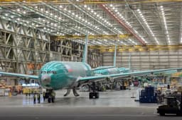 The interior of the Boeing Everett Factory: two unpainted green-primer 777X widebody fuselages with tails and wings attached, parked nose-forward on the assembly floor under a lit steel roof truss with overhead cranes, with tooling and small figures of workers in high-visibility vests alongside.