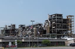 A daylight view of an industrial chemical plant: multi-story steel process units dense with piping, ducting, and stair towers rising behind a low windowless building, a chain-link perimeter fence, and an empty employee parking lot in the foreground.