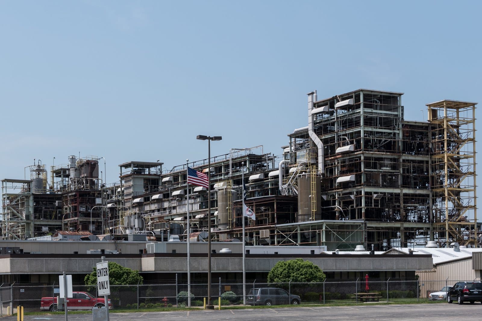 A daylight view of an industrial chemical plant: multi-story steel process units dense with piping, ducting, and stair towers rising behind a low windowless building, a chain-link perimeter fence, and an empty employee parking lot in the foreground.