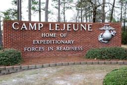 Brick welcome sign at the Holcomb Gate entrance to Marine Corps Base Camp Lejeune reading 'CAMP LEJEUNE — Home of Expeditionary Forces in Readiness,' with the Marine Corps Eagle, Globe, and Anchor emblem mounted on the right side