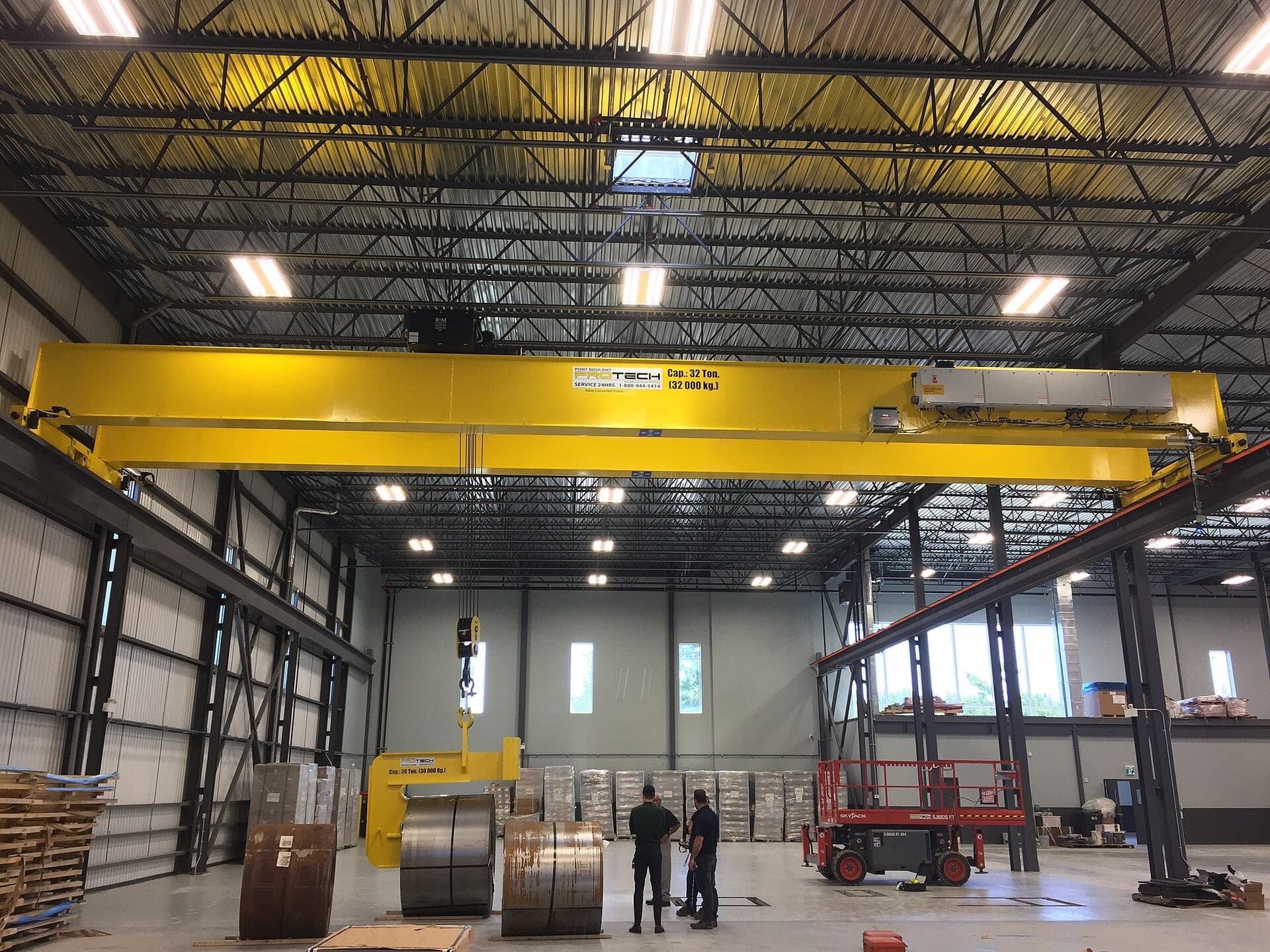 A yellow overhead bridge crane spanning the interior of an industrial fabrication facility, with a large metal coil resting on the floor below it.