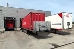 Tractor-trailers and a shipping container parked on the apron beside a warehouse loading dock, with an empty dock bay and dock shelter along the building wall.