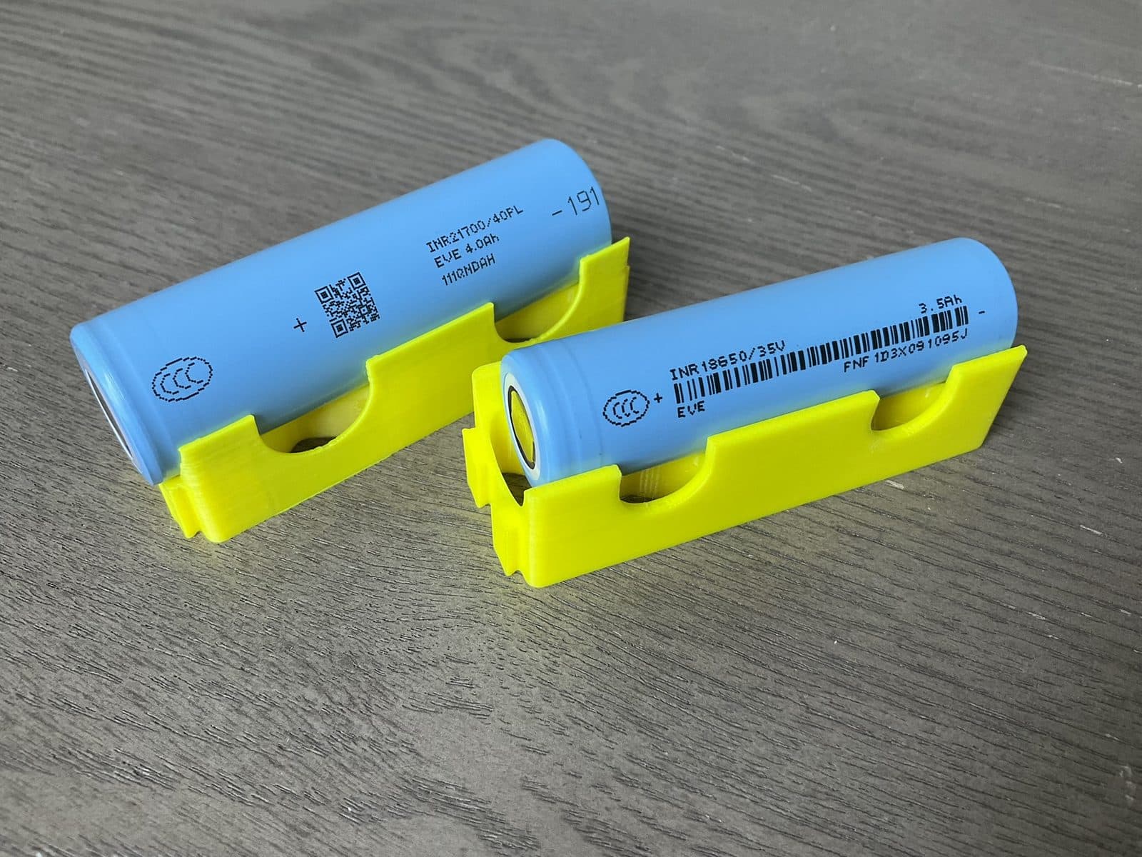 Two cylindrical lithium-ion battery cells (18650 and 21700 format) resting in yellow holders on a wood surface, with generic cell markings and no consumer-product branding.