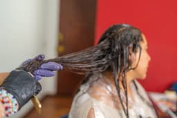 A gloved stylist works a chemical hair relaxer through a client's hair