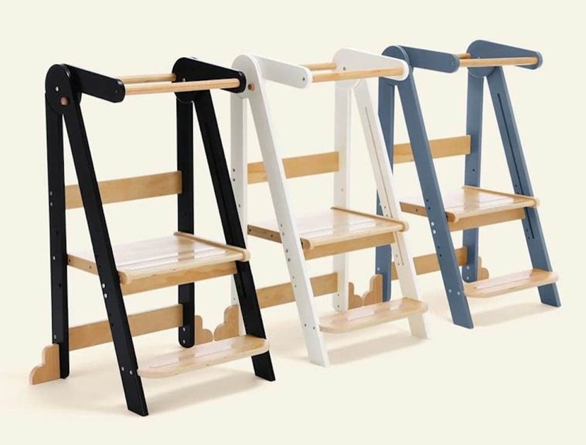 Three HARPPA Nordi foldable toddler tower stools in black, white, and blue finishes with natural-wood steps, platforms, and safety bars, standing side by side against a plain background.
