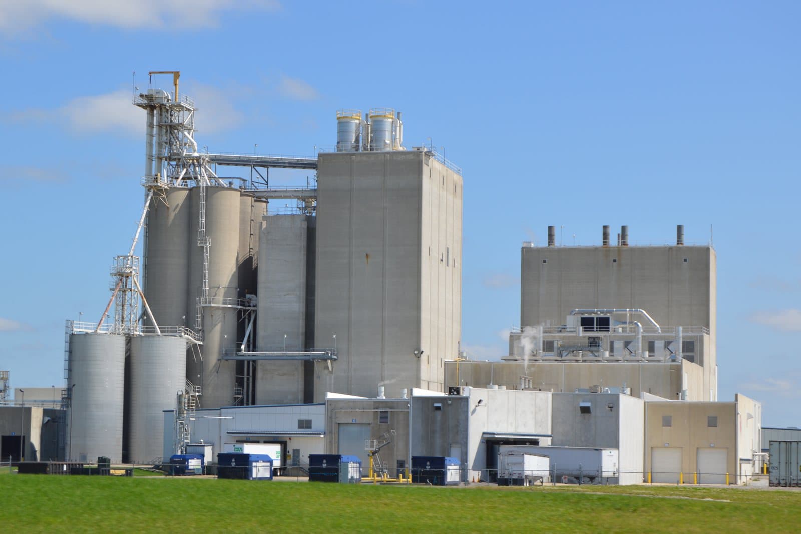 A pet food manufacturing plant seen from a grass field: concrete process towers and storage silos linked by elevated conveyors, a taller windowless production block with roof stacks, and truck bays with a parked trailer along the ground floor.