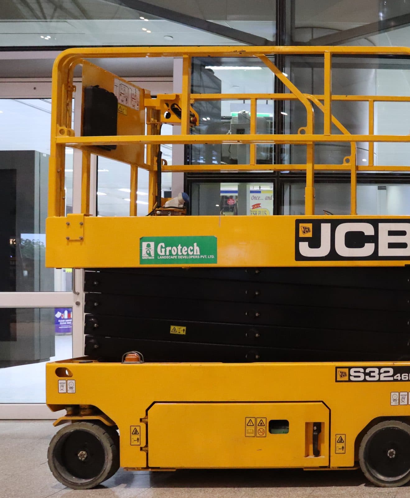 A yellow JCB scissor lift, a self-propelled aerial work platform, standing fully lowered on its wheels inside a glass-walled facility.