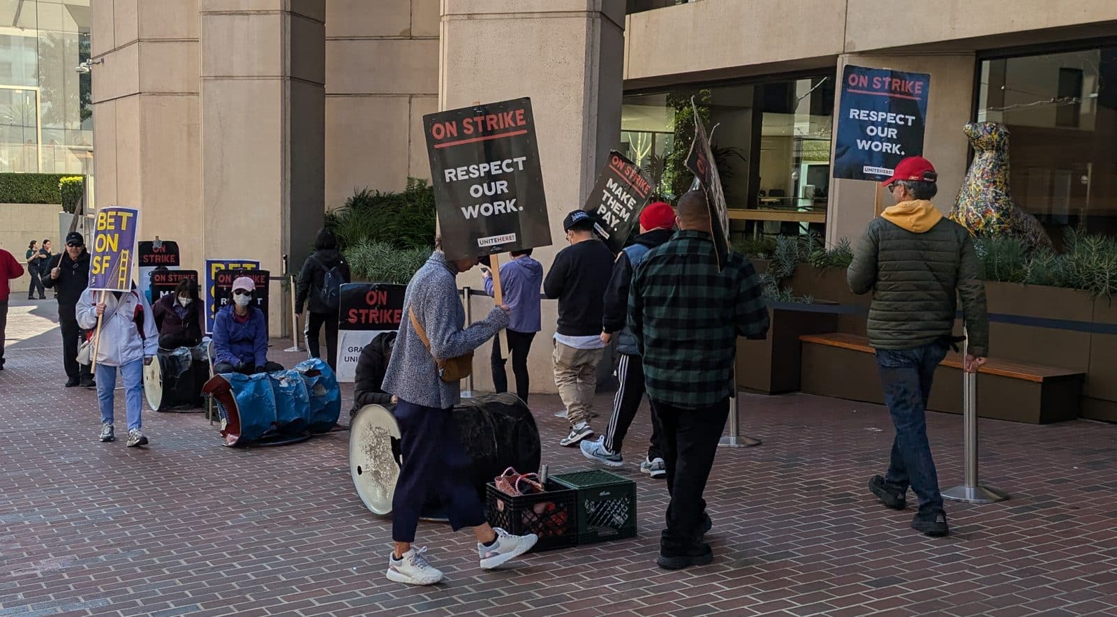 Hotel workers walk a picket line on a brick plaza outside a hotel, carrying black 'On Strike — Respect Our Work' signs marked UNITE HERE, most seen from behind.