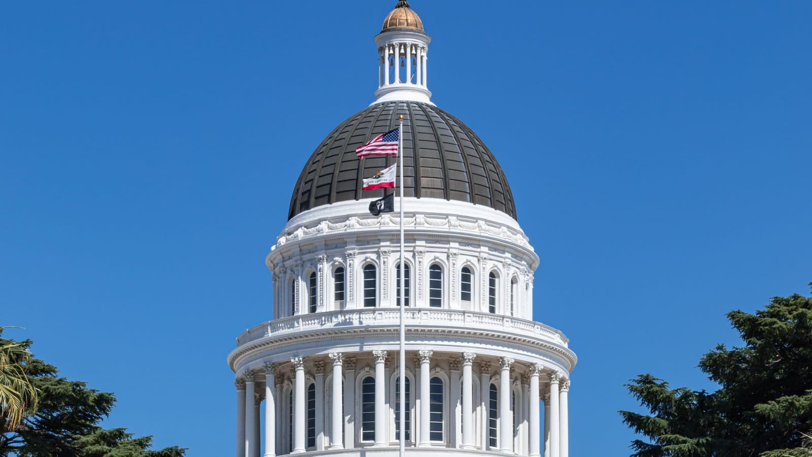 The white dome and columned front of the California State Capitol in Sacramento, flying the U.S. and California flags against a clear blue sky, framed by trees.