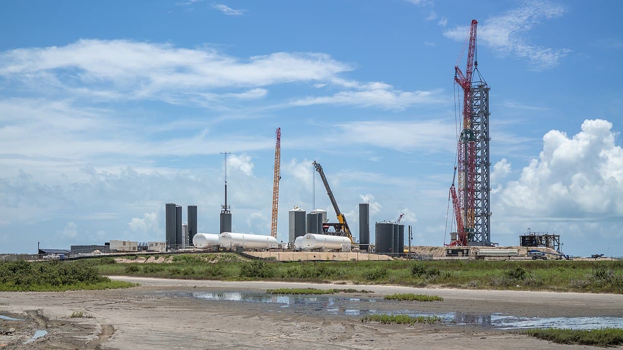 The SpaceX Starbase launch site at Boca Chica, South Texas, showing a launch tower under construction, cranes and storage tanks across coastal marsh terrain.