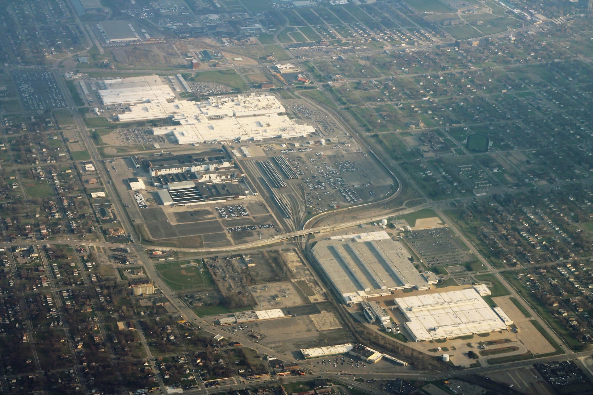 Aerial view of a large automotive assembly plant complex in Detroit, with expansive rooftops, parking lots filled with vehicles, and rail lines running through an urban industrial area.