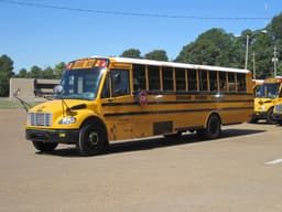 A yellow Durham School Services school bus parked at a bus depot, with more school buses of the same fleet lined up behind it under a clear sky.