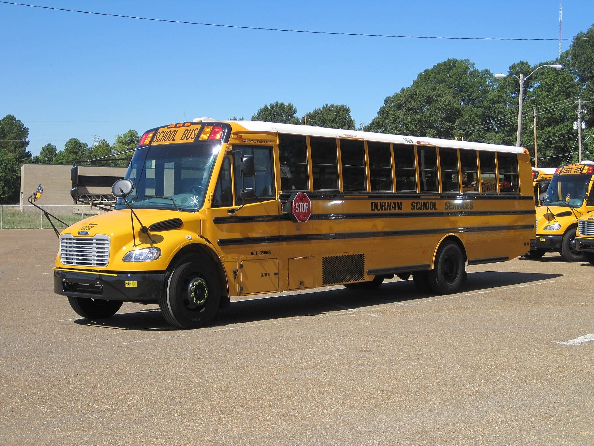 A yellow Durham School Services school bus parked at a bus depot, with more school buses of the same fleet lined up behind it under a clear sky.