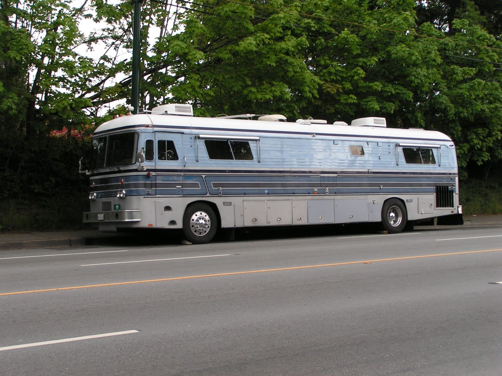 A silver bus-style Class A motorhome parked alongside a road in daytime, with trees behind it and no people in frame.