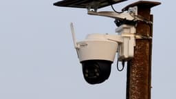 A white solar-powered outdoor pan-and-tilt security camera with an attached solar panel, dome lens, and antenna, mounted outdoors against a daytime sky, with no people in frame.